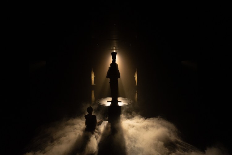 A seated woman gazes at the statue of Artemis in silhouette as clouds of fog billow on the floor